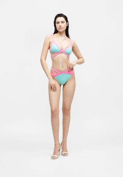 Γυναίκα που φοράει μπλε και ροζ μπικίνι με ανοίγματα, και λευκά σανδάλια με ψηλό τακούνι, σε απλό λευκό φόντο.