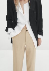 Musta kaksirivinen bleiseri valkoisen pellavapaidan päällä, yhdistettynä beigen värisiin räätälöityihin housuihin, joissa on etutasku ja napin yksityiskohta.