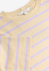 T-shirt beige à col rond avec des rayures horizontales violet clair et une manche repliée, montrant la texture du tissu et les détails de la couture.