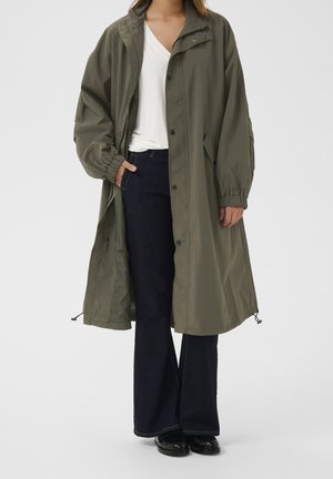Femme portant une longue veste oversize vert olive sur un top blanc à col en V, jean large foncé et chaussures noires, debout avec une main dans la poche.