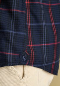 Granatowa kraciasta koszula z czerwonymi i niebieskimi paskami, z naszywką z logo na dolnym brzegu. Nosi się ją na jasnobeżowych spodniach z korduroy.