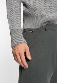 Grau gestrickter Pullover mit gerippten Bündchen, kombiniert mit dunkelgrauen Hosen, die ein sichtbares "BOSS"-Label auf der Tasche haben.