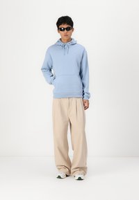 Hellblaues Hoodie mit Kordelzug, Kängurutasche, kombiniert mit beigen weit geschnittenen Hosen und weißen Sneakers mit grünen Akzenten. Modell trägt Sonnenbrille.