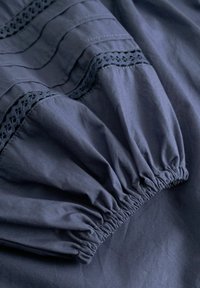 Tmavě modrá tkanina s elastickým lemem a ozdobnými krajkovými vsadkami, zobrazující nařasený a přišitý detail na rukávu oděvu.