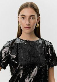 Sort pailletetop med rund hals og korte ærmer, med en skinnende overflade og firkantet pailletmønster. Guld hoops øreringe medfølger.