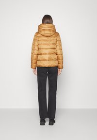 Puffjakke i varm karamelfarve med horisontal quilting, hætte og afslappet pasform, parret med sorte lige ben bukser.