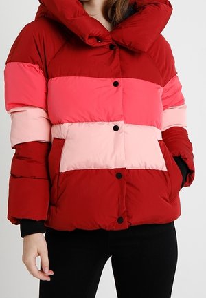 Femme portant une doudoune à blocs de couleurs avec des rayures horizontales rouges, rose foncé et rose clair, boutons sur le devant et poches latérales.
