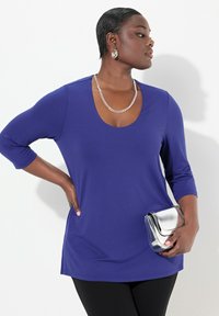 Maglietta lunga viola con scollatura a barca, abbinata a leggins neri e una borsa metallica argento. Tessuto con texture e finitura liscia.
