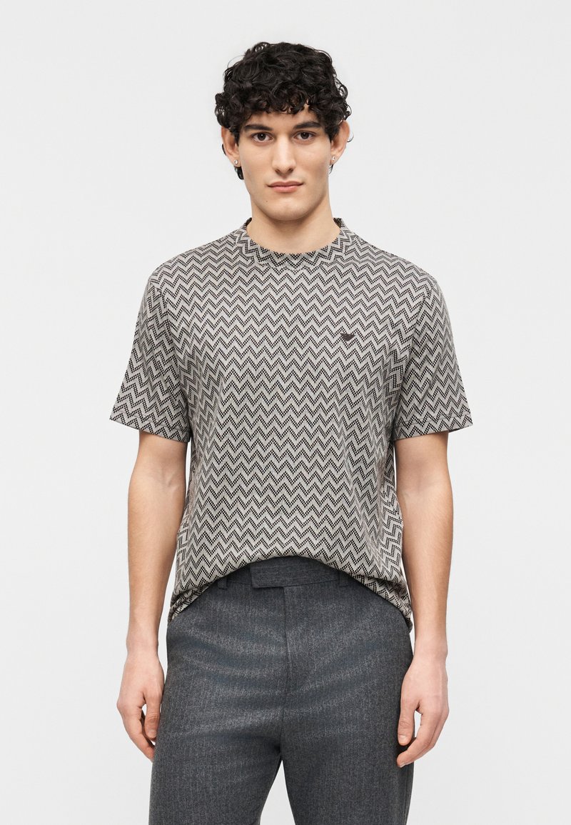 Νεαρός άντρας με κοντά σγουρά μαλλιά, που φοράει πουκάμισο με σύντομο μανίκι και ζιγκ ζαγκ μοτίβο, και σκούρο γκρι παντελόνι, στέκεται μπροστά σε απλό φόντο.