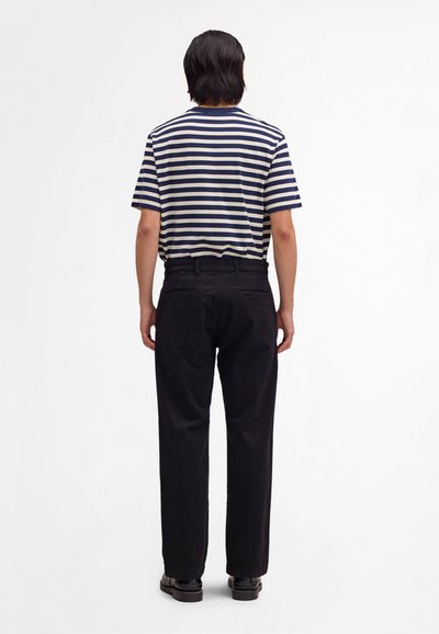 Person mit mittellangen dunklen Haaren steht mit dem Rücken zur Kamera, trägt ein kurzärmeliges Shirt mit marineblau-weißen Streifen, schwarze Hose und schwarze Schuhe.