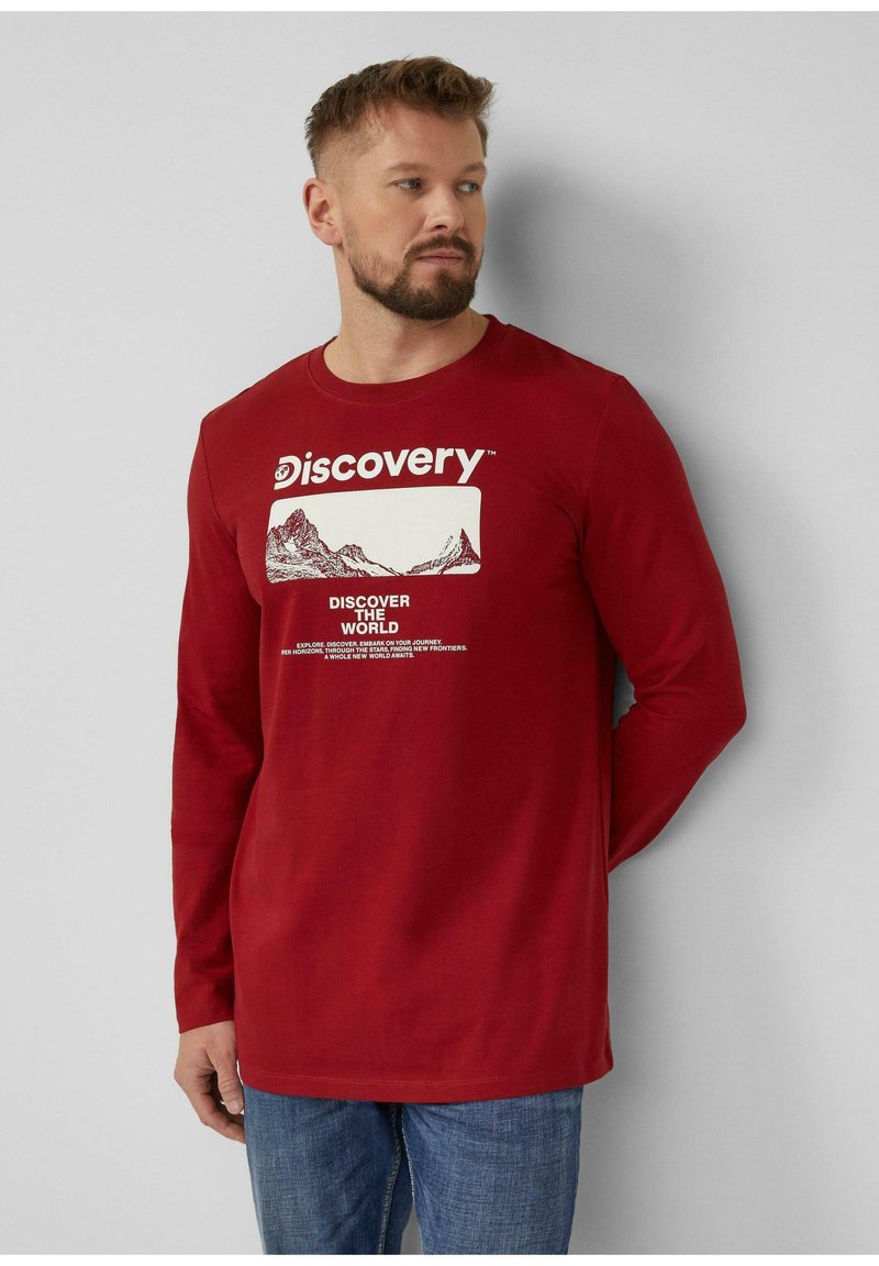 Homme à la barbe portant une chemise rouge à manches longues avec un graphique de montagne et le texte blanc "Discovery", debout devant un mur gris, regardant vers la droite.