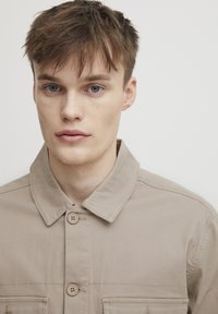 Beige cotton jacket with a collared neckline, front buttons, and chest pockets. Smooth texture and casual design.