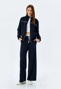Marinblå träningsoverall i mjukt tyg med en zip-up jacka och vita detaljer samt avslappnade byxor. Vita sneakers kompletterar outfiten.