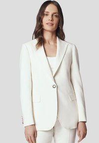 Biała marynarka dopasowana z klapami, z zapięciem na pojedynczy guzik, kieszeniami z przodu i gładkim, usztywnionym materiałem. W połączeniu z białą bluzką.
