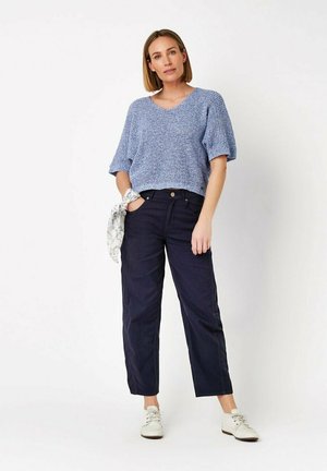 Donna in piedi con la mano in tasca, che indossa un maglione a maniche corte azzurro chiaro con texture, pantaloni scuri a gamba larga, sneakers bianche e una sciarpa a fantasia al polso.