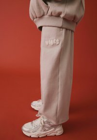 Jasnoróżowe dresy z haftem "Good Vibes" w puszystych literach z boku, w połączeniu z błyszczącymi białymi sneakersami na czerwonym tle.