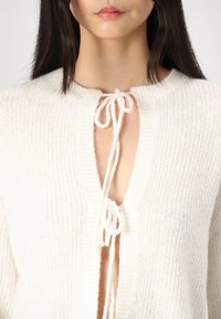White knit cardigan featuring a V-neckline, tie closure at the center, and ribbed texture throughout. Soft fabric with a lightweight design.