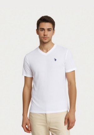 Young man wearing a plain white V-neck T-shirt with a small blue embroidered logo on the left chest and beige pants.