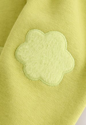 Limoengroen stof met een pluizige, lichtgroene bloemvormige patch; een gestikte rand omlijnt het bloemmotief. Zachte textuur en levendige kleur.