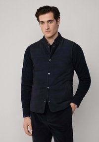 Mornarska prsluk s kapicom, ukrašen dugmadi u boji, i bočnim džepovima, složen preko teksturirane mornarske pletene majice. Glatka završna obrada tkanine.