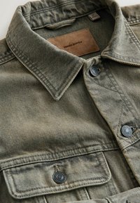 Olīvu zaļas krāsas džinsa krekls ar asu apkakli, dubultām krūšu kabatām, melniem pogām un brūnas ādas etiķeti ar uzrakstu "DENIM SUPPLY".