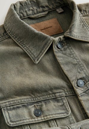 Olivovo zelená denimová košeľa s ostrým golierom, dvojitými náprsnými vreckami, čiernymi gombíkmi a béžovou koženou štítkom s nápisom "DENIM SUPPLY".