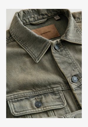 Olīvu zaļas krāsas džinsa krekls ar asu apkakli, dubultām krūšu kabatām, melniem pogām un brūnas ādas etiķeti ar uzrakstu "DENIM SUPPLY".