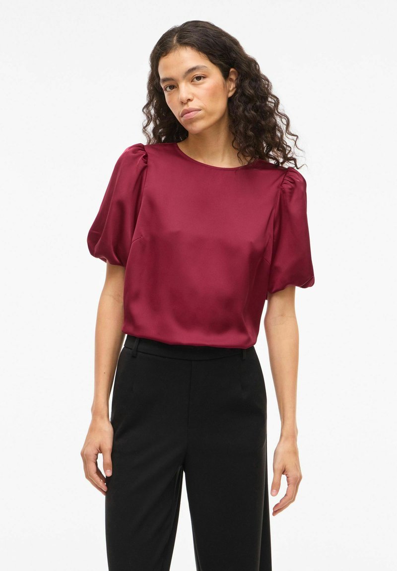 Blouse en satin bordeaux avec manches courtes bouffantes, encolure ronde et texture lisse, associée à un pantalon noir.