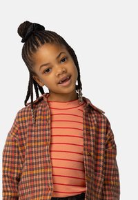 Young girl with braided hair and beads, wearing an orange striped shirt under a multicolored houndstooth jacket, tilting head slightly.