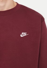 Vínová mikina vyrobená z bavlneného zloženia, s okrúhlym výstrihom a malým bielym logom Nike na hrudi. Hladká textúra, klasický strih.