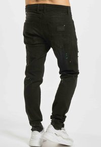 Črne jeans hlače iz denima z ozkim krojem, obrabljenimi detajli, škropljenim zelenim barvami in zadnjimi žepi. V kombinaciji z belimi teniskami.