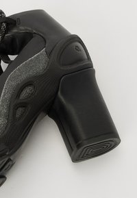 Svart helfot som har en teksturert overflate, med en glitrende grå detalj og en kraftig hæl. Snøringer og utskårne detaljer forbedrer designet.