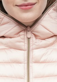 Lyserød, fyldig jakke med quiltet struktur, der har en guldfarvet lynlås og høj krave. Lynlåstrækker i blød lyserød. Nærbillede af halsudskæringen.