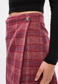 Sarkans pārbaudīts šorti, izgatavoti no teksturēta auduma, ar sudraba pogu akcentu un apvītu dizainu. Redzamās horizontālās un vertikālās raksta līnijas.