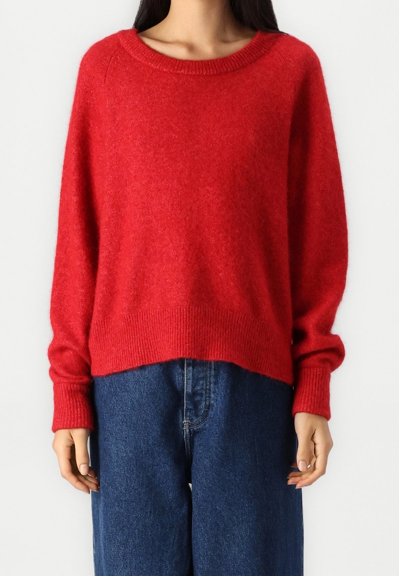 Femme portant un pull en maille rouge ample à manches longues et un jean taille haute bleu foncé, debout devant un fond blanc.