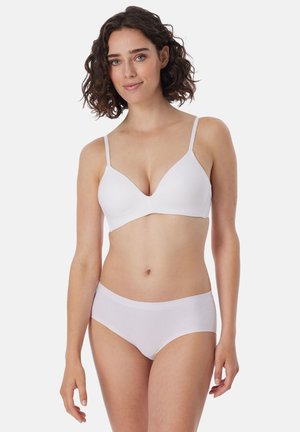 Frau mit lockigem Haar, die einen weißen BH mit dünnen Trägern und passende weiße nahtlose Unterwäsche trägt, steht vor einem schlichten Hintergrund.