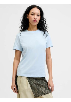 Göndör fekete hajú nő világoskék rövid ujjú pólóban és bézs szoknyában, fekete csipkés részlettel, egyszínű háttér előtt állva.