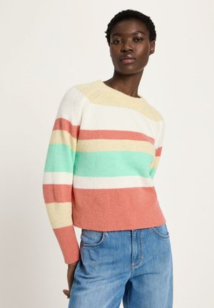 Frau trägt einen mehrfarbigen gestreiften Pullover mit pastellgelben, korallenroten, mintfarbenen und weißen Streifen, kombiniert mit blauen Jeans, und steht vor einem einfachen Hintergrund.