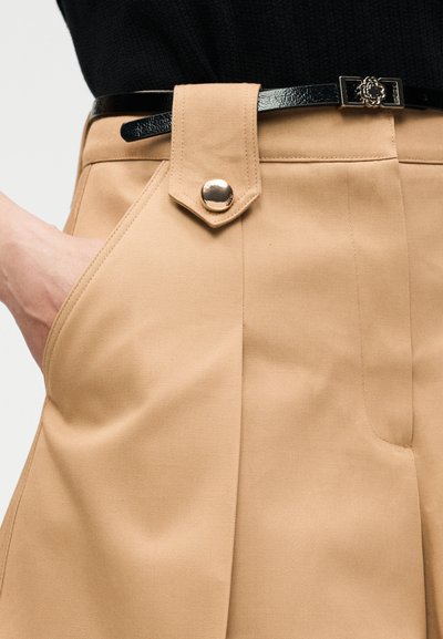 Μπεζ παντελόνι ψηλόμεσο με πλαϊνή τσέπη, θηλιά με κουμπί και λεπτή μαύρη ζώνη με αγκράφα σε σχήμα λουλουδιού.
