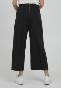 Schwarze weitgeschnittene Hose aus glattem Stoff mit einem Reißverschluss auf der Rückseite. Kombiniert mit weißen Sportschuhen hat das Outfit ein minimalistisches Design.