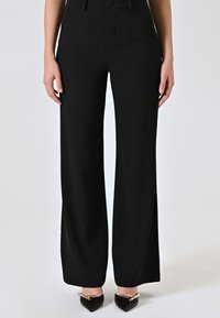 Pantaloni neri a gamba larga realizzati in tessuto liscio con una finitura pulita, dotati di chiusura frontale e passanti per cintura, abbinati a scarpe nere con punta.
