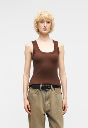 Άτομο με κοντά ξανθά σγουρά μαλλιά που φοράει καφέ αμάνικο τοπ μέσα σε χαλαρό μπεζ παντελόνι με μαύρη ζώνη, στέκεται μπροστά σε απλό φόντο.