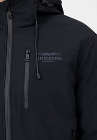 Zwarte capuchonjack met een gladde textuur, ritszakken, verstelbare trekkoorden en een subtiel logo met vleugels op de borst.