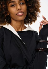 Zwarte top met een witte kraag, met een zachte textuur. Model versierd met bruine, ronde armbanden en oorringen.