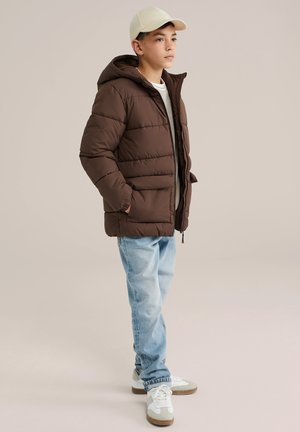 Veste matelassée marron avec capuche, présentant un design matelassé et des poches latérales, portée par-dessus une chemise grise et assortie à un jean bleu clair et des baskets blanches.