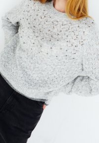 Femme portant un pull en maille gris clair texturé avec de petites perles, associé à un pantalon noir taille haute, debout les mains dans les poches.