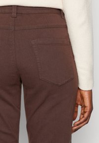 Pantaloni in denim marrone con una tasca posteriore, caratterizzati da cuciture visibili, una texture liscia e un design aderente. Un maglione leggero è anche parzialmente visibile.