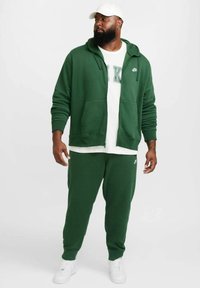 Grön Nike zip-up hoodie med framfickor, ihopparad med matchande sweatpants. Vit t-shirt under med "NIKE" text. Vita sneakers.