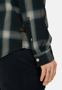 Koszula w kratę z długim rękawem w kolorze granatowym i beżowym. Charakteryzuje się subtelną fakturą, zaokrąglonym dołem i mankietami na guziki. Metka przyszyta na bocznym szwie.