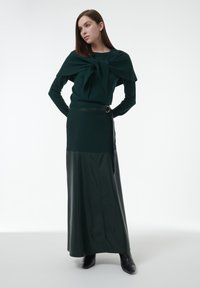 Donkergroene gebreide trui met een strikdetail, gecombineerd met een donkergroene leren maxirok, afgewerkt met een riem en zwarte enkellaarsjes.